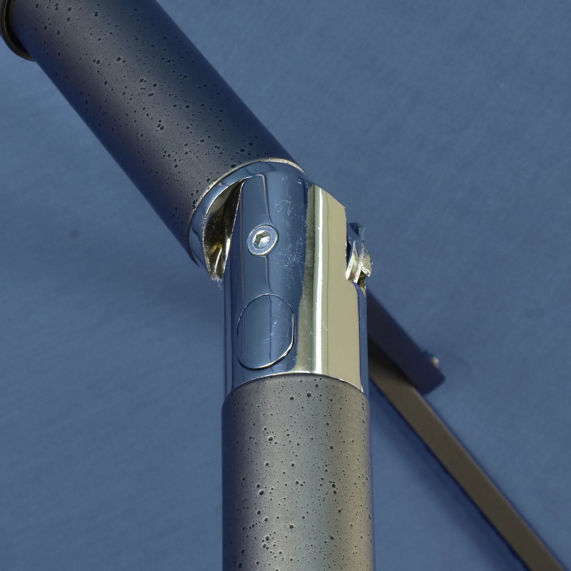 Close-up image of the simple push-button tilt mechanism on the Pacific Trail Series Patio Umbrella by California Umbrella. This detailed view captures the shiny, metallic button set against the textured aluminum pole, illustrating the ease of adjusting the umbrella's angle for optimal shade coverage. The functionality and strategic placement of the button ensure that users can easily modify the canopy's position to provide continuous comfort throughout the day in various outdoor settings.