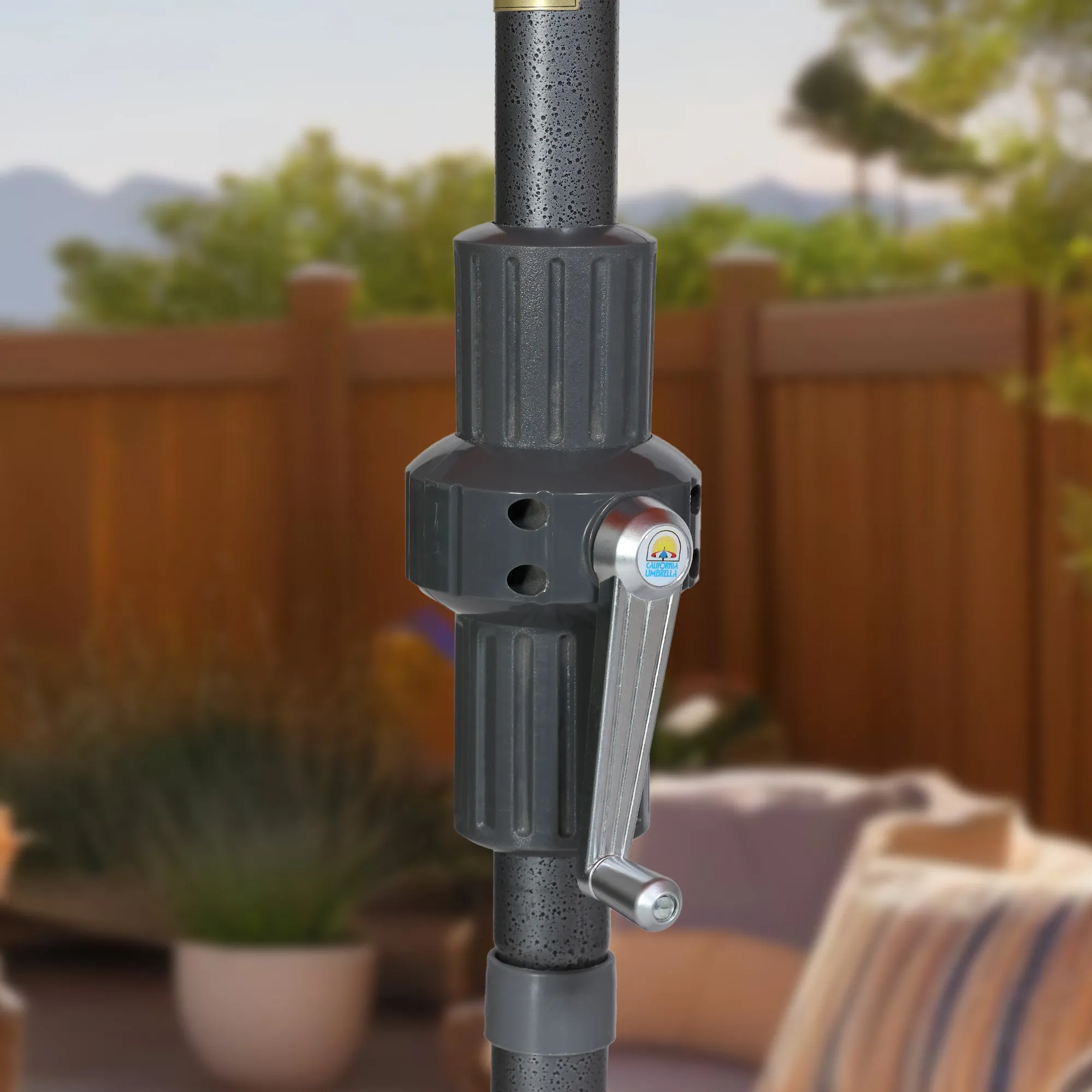 Close-up image of the easy crank lift mechanism on the Sun Master Series Patio Umbrella by California Umbrella, set against a scenic outdoor backdrop. This detailed view displays the sleek, gray crank handle that simplifies the opening and closing of the umbrella. Designed for ease and efficiency, the crank lift is integral to the umbrella's user-friendly functionality, allowing for quick adjustments with minimal effort. The backdrop features a relaxed patio setting, enhancing the outdoor leisure appeal of the Sun Master Series.