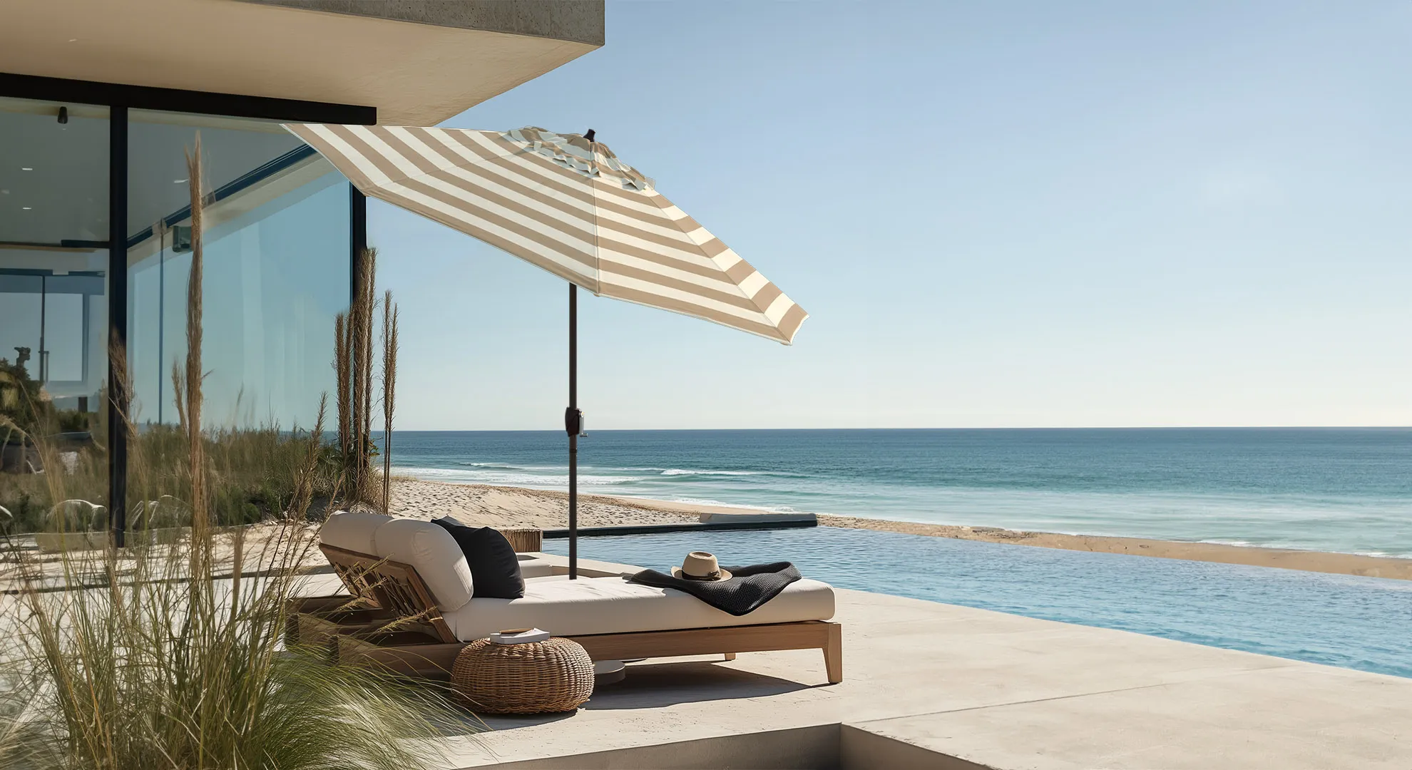 Hero banner image of the Vento Series Residential Patio Umbrella by California Umbrella, set against a serene coastal backdrop. This umbrella, crafted with high-quality Sunbrella fabric, features a stylish striped design that complements the luxurious outdoor setting. Positioned beside a modern glass house overlooking the beach, it exemplifies sophisticated coastal living. The umbrella provides ample shade over a contemporary outdoor lounger, enhancing the residential patio experience with its durable and elegant construction. Ideal for anyone seeking a blend of style, comfort, and long-lasting performance under the California sun.